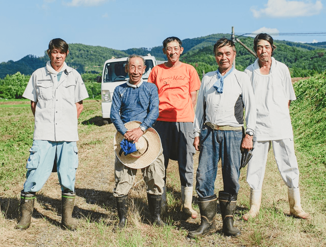 畑の前で並んで立つ5人の農家の男性たち（背景に軽トラックと山）