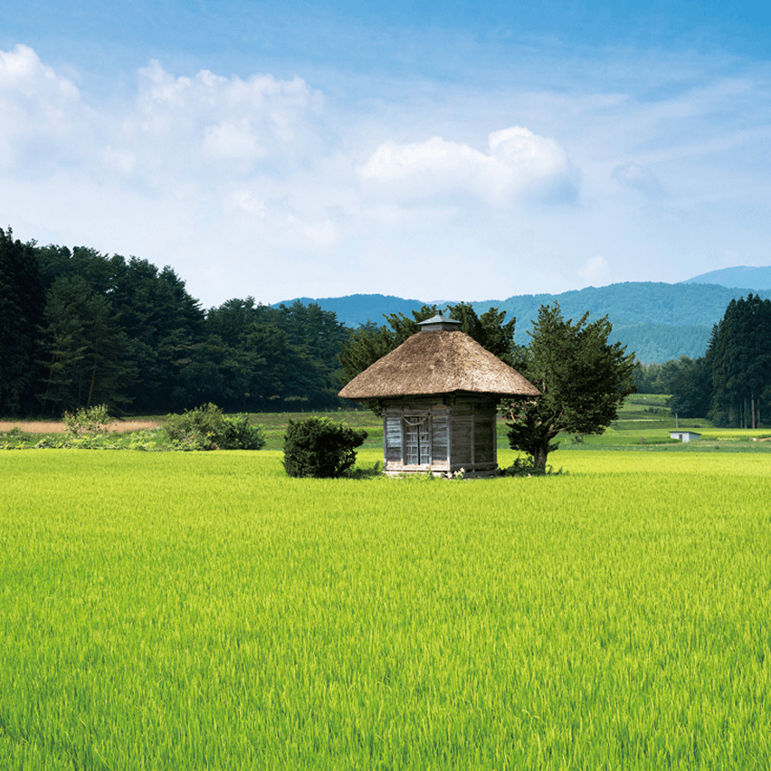 青空の下、緑の田んぼの中に茅葺き屋根の小屋が建つ遠野ののどかな田園風景（背景に森と山並み）